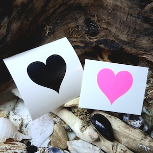 May include: Two heart-shaped stickers. One sticker has a black heart on a white background. The other sticker has a pink heart on a white background. The stickers are displayed on a bed of seashells and driftwood.