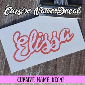 May include: A white decal with the name "Elissa" in a red cursive font. The decal is on a white background, with the text "Cursive Name Decal" above and below the name. The decal is held by a metal clip.