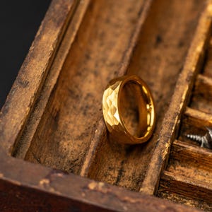 May include: A gold ring with a textured, faceted surface sits in a wooden box. The ring's geometric design reflects light, highlighting its metallic sheen. The box has a warm, brown hue, providing a contrasting backdrop for the jewellery.