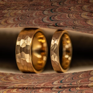 Op de afbeelding: Twee gouden trouwringen met een gehammerde textuur. De ringen liggen op een boek met een marmeren omslag.