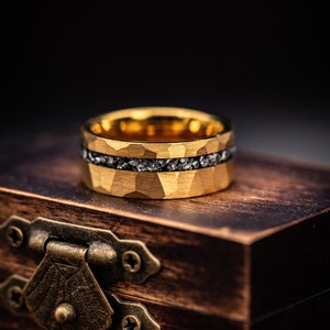 Men's Gold tungsten engagement ring with hammered finish and silver meteorite inlay laying on its side on top of a walnut ring box