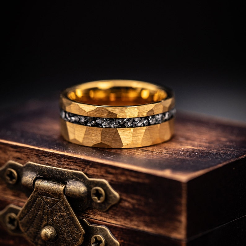 Men's Gold tungsten engagement ring with hammered finish and silver meteorite inlay laying on its side on top of a walnut ring box