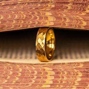 May include: A gold ring with a textured, geometric pattern stands upright between the pages of a book. The ring's surface reflects light, and the book's pages have a patterned design in shades of red and brown.