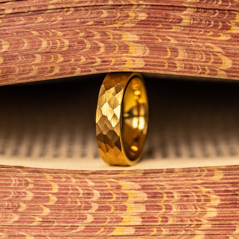 May include: A gold ring with a textured, geometric pattern stands upright between the pages of a book. The ring's surface reflects light, and the book's pages have a patterned design in shades of red and brown.
