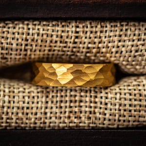 May include: A gold-coloured ring with a textured, geometric pattern. The ring is displayed inside a dark wooden box lined with a woven, tan fabric. The ring's surface reflects light, highlighting the carved design.