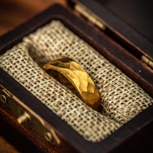 May include: A gold ring with a textured, faceted surface sits in a dark wooden box lined with tan fabric. The ring's design features geometric shapes, creating a unique appearance. The box is open, revealing the ring. The ring is a stylish piece of jewellery.