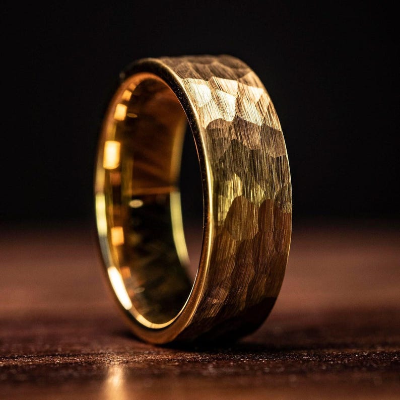 Men's Yellow gold tungsten wedding band with hammered finish standing upright on oak table