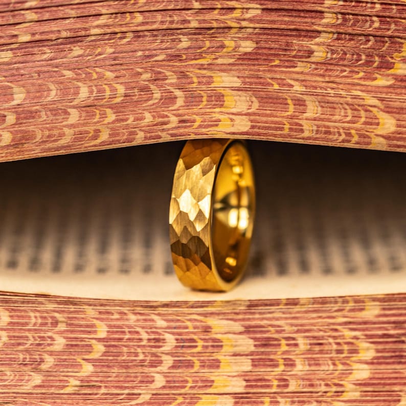 May include: A gold ring with a textured, faceted surface stands upright between the pages of a vintage book. The ring's design catches the light, showcasing its intricate details. The book's pages are aged with a reddish-brown and yellow pattern.