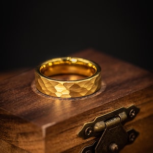 May include: A gold ring with a hammered finish. The ring sits on a wooden box with a brass clasp. The ring's surface reflects light, emphasizing the textured design. The ring is a piece of jewellery.