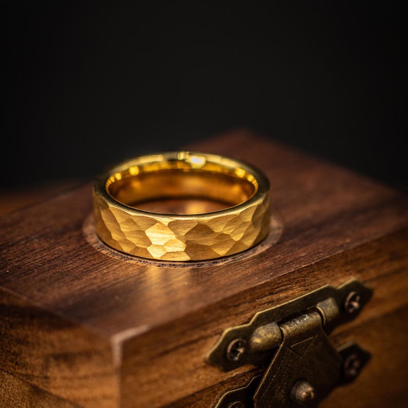 May include: A gold ring with a hammered finish. The ring sits on a wooden box with a brass clasp. The ring's surface reflects light, emphasizing the textured design. The ring is a piece of jewellery.