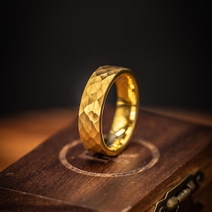 May include: A gold ring with a textured, hammered finish. The ring is displayed on a wooden box against a dark backdrop. The ring's surface is faceted, giving it a distinctive look. The ring is a piece of jewellery, suitable for everyday wear or special occasions.