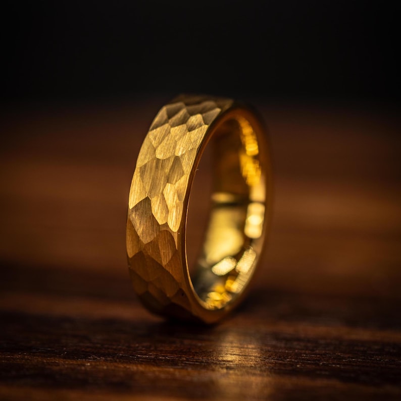 May include: A gold ring with a textured, hammered finish. The ring has a geometric pattern, reflecting light. The ring is a solid band. The ring is set against a dark background, highlighting its metallic sheen. This ring is a piece of jewellery.