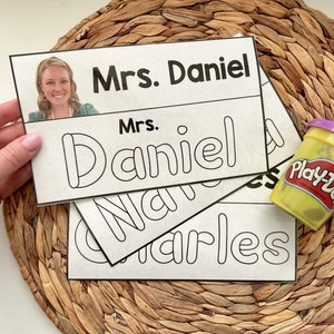 以下が含まれることがあります： 黒い文字が書かれた白いカードが3枚。一番上のカードには「Mrs. Daniel」と書かれている。真ん中のカードには「Mrs. Daniel」と、名前がバブルフォントで書かれている。一番下のカードには「Natalie Charles」と、名前がバブルフォントで書かれている。画像の右下には、小さな黄色のプレイ・ドーの容器がある。