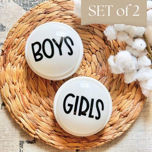 May include: Two white, round decorative objects with the words "BOYS" and "GIRLS" in black lettering. The objects are set on a woven, circular placemat. The words "SET of 2" are in the upper right corner.