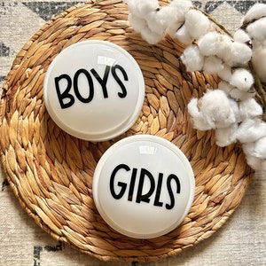K&ouml;nnte beinhalten: Zwei wei&szlig;e runde Leuchtzeichen mit schwarzem Text. Das Schild links sagt "BOYS" und das Schild rechts sagt "GIRLS".