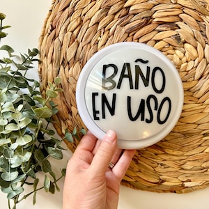 K&ouml;nnte beinhalten: Ein wei&szlig;es rundes Schild mit schwarzem Text, der "Ba&ntilde;o en uso" lautet, was im Deutschen "Bad belegt" bedeutet. Das Schild befindet sich auf einem geflochtenen Strohhintergrund.