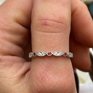 May include: A delicate silver band ring with a row of small, clear gemstones and red accents.