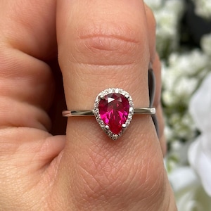 May include: A pear-shaped ruby ring with a halo of smaller diamonds set in a silver band.