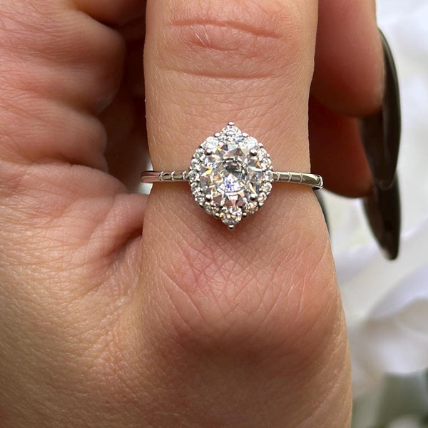 Op de afbeelding: Een zilveren ring met een grote, ronde, heldere diamant omringd door kleinere diamanten. De ring zit aan een vinger.