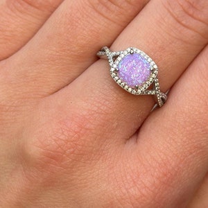 May include: A silver ring with a large, round, purple opal gemstone surrounded by a halo of smaller clear stones. The ring has a twisted band.