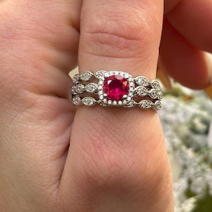 May include: A three-piece ring set featuring a red gemstone surrounded by smaller clear stones. The ring is on a finger with white nail polish.