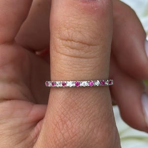 May include: A delicate ring with alternating white and pink gemstones. The ring is set on a finger, showcasing the intricate design and sparkle of the gemstones. The ring is set against a backdrop of white and blue flowers.