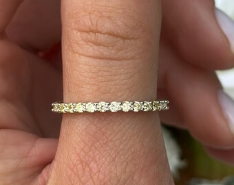 May include: A gold ring with a row of small, clear gemstones. The ring is on a person's finger, with a blurred floral background. The ring's band is thin, and the gemstones are evenly spaced around the top.