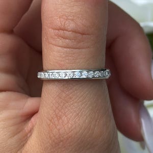 May include: A silver band ring set with small, clear gemstones. The ring is displayed against a backdrop of white flowers and green leaves. The ring is on a finger with long, light-coloured nails.