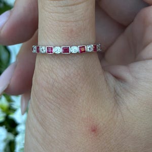 May include: A silver ring with alternating red and clear stones. The ring is worn on a finger, with a blurred floral background. The ring's stones are rectangular and round, creating a delicate and elegant design.
