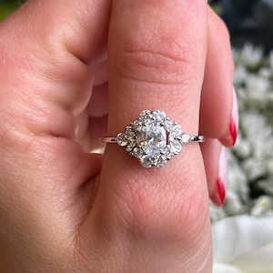 May include: A silver ring with a large oval-shaped center stone surrounded by smaller diamonds. The ring is set in a floral design with delicate details.