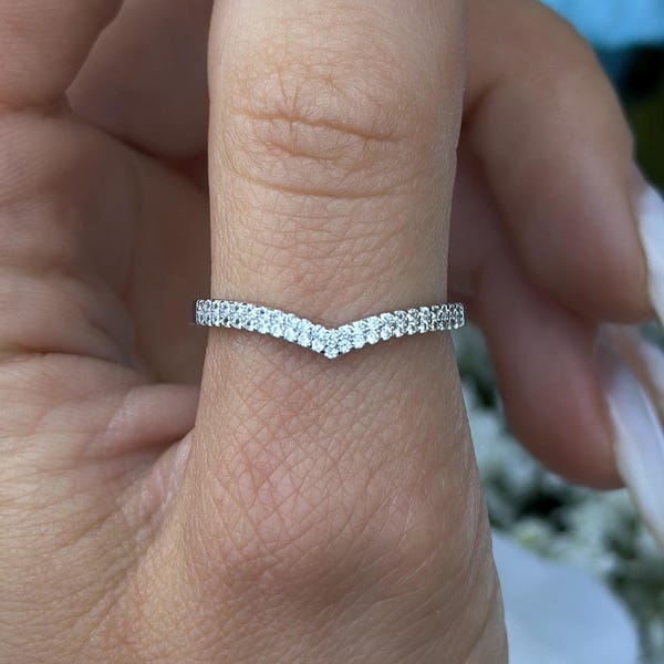 Op de afbeelding: Een zilveren V-vormige ring met kleine, heldere edelstenen langs de band. De ring zit om een vinger, met een vage bloemenachtergrond van witte en blauwe bloemen.