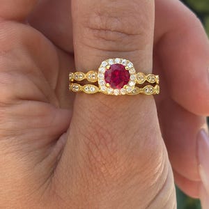May include: A gold ring set featuring a ruby center stone surrounded by small white stones. The ring is held up against a blurred floral background. The set includes a matching band with a repeating pattern of small white stones.