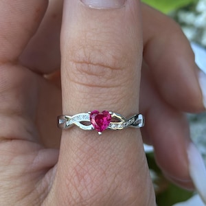 May include: A silver ring with a heart-shaped, red gemstone at the center. The ring features a twisted band design with small, clear accent stones. The ring is shown on a finger against a backdrop of white and blue flowers.