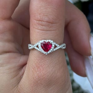 May include: A silver ring with a heart-shaped, ruby-red center stone, surrounded by a halo of smaller, clear stones. The ring's band features a delicate, twisted design. The ring is displayed against a backdrop of white and blue flowers.