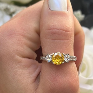 May include: A silver ring with a large yellow center stone and two smaller clear stones on either side. The ring is set with small clear stones around the band.