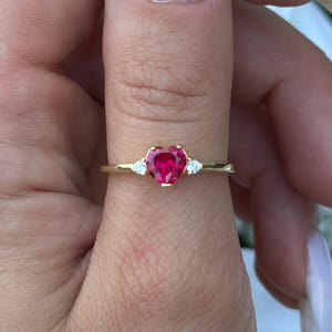 May include: A gold ring featuring a heart-shaped, bright red gemstone. Two small, clear gemstones flank the heart-shaped stone. The ring is set on a slender gold band. The ring is shown on a hand with long, white fingernails.