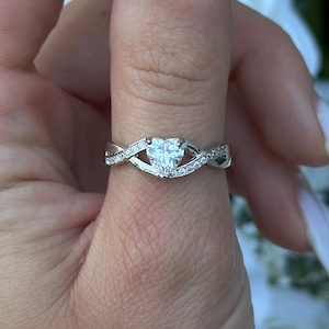 May include: A silver ring with a heart-shaped diamond centre stone. The ring features a twisted band design with smaller accent stones. The ring is shown on a finger with long, light-coloured nails, with a blurred floral background.