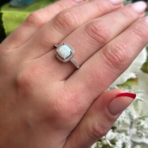 May include: A silver ring with a white opal gemstone surrounded by smaller clear gemstones. The ring is on a finger with white nail polish.
