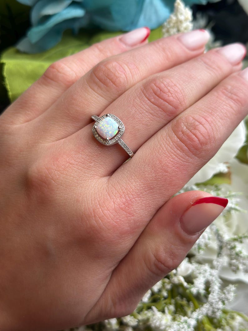 May include: A silver ring with a white opal gemstone surrounded by smaller clear gemstones. The ring is on a finger with white nail polish.