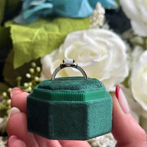 May include: A silver ring with a white opal gemstone set in a square bezel. The ring is in a green velvet ring box.