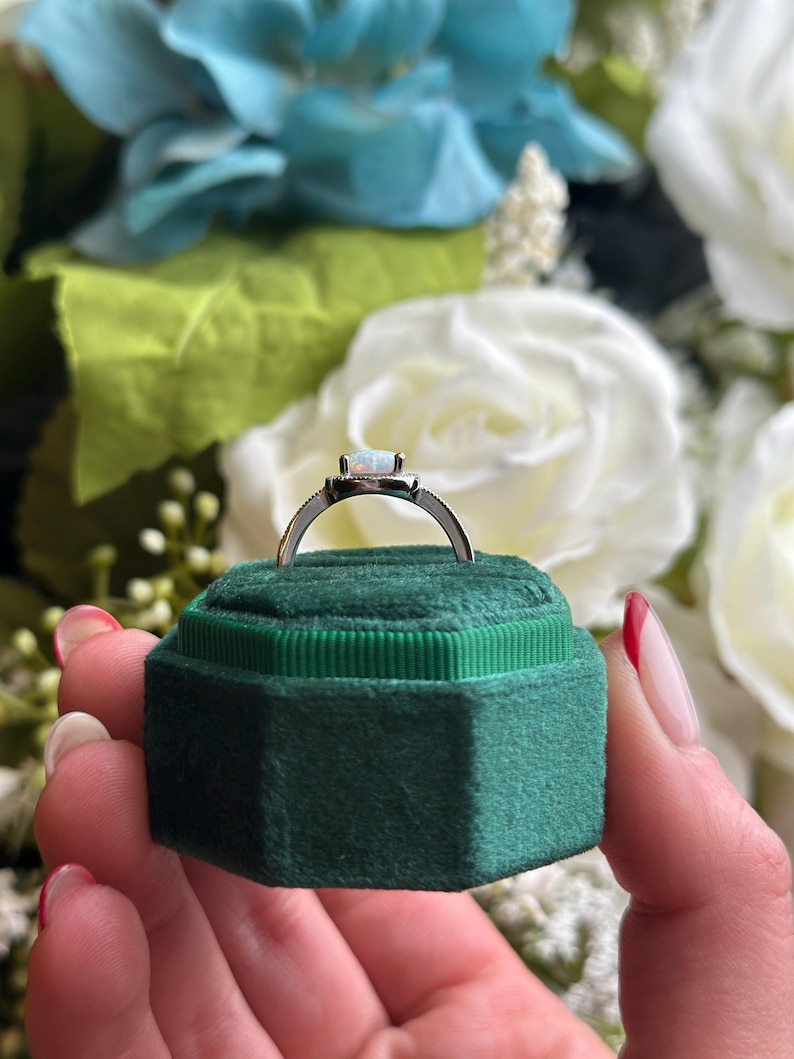 May include: A silver ring with a white opal gemstone set in a square bezel. The ring is in a green velvet ring box.
