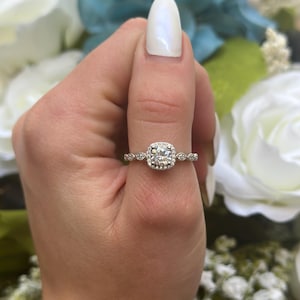 Peut inclure: Une bague en argent avec une pierre centrale ronde et transparente entourée de pierres plus petites. La bague est sertie sur une bande blanche.