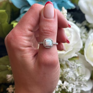 May include: A silver ring with a white opal gemstone surrounded by smaller clear gemstones. The ring is on a finger with red nail polish.