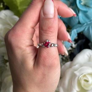 Peut inclure: Une bague en argent avec une pierre rouge en forme de cœur et une petite pierre transparente. La bague est de style nœud celtique.