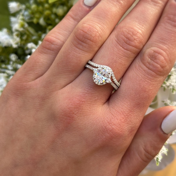 Op de afbeelding: Een zilveren verlovingsring met een grote ovale middelste steen omringd door kleinere diamanten. De ring zit op een vinger met witte nagellak.