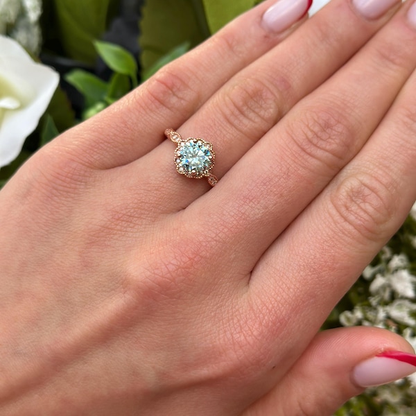 Op de afbeelding: Een verlovingsring van roségoud met een grote, ronde, lichtblauwe edelsteen omringd door kleinere diamanten. De ring zit aan een vinger met lichtroze nagellak.