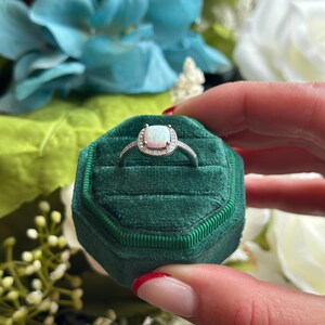May include: A silver ring with a white opal gemstone surrounded by smaller clear gemstones. The ring is in a green velvet ring box.