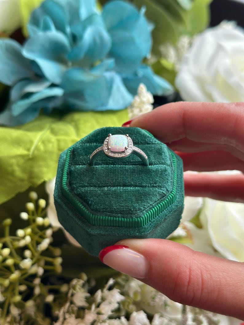 May include: A silver ring with a white opal gemstone surrounded by smaller clear gemstones. The ring is in a green velvet ring box.