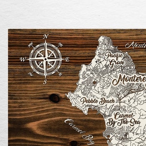 May include: A wooden map of Monterey, California, with a compass rose in the upper left corner. The map shows the city's streets and landmarks, including Carmel-by-the-Sea, Pebble Beach, and Del Ray Oaks. The text "Papier Blanc" is printed in the lower left corner.
