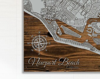May include: A wooden wall art piece featuring a detailed map of Newport Beach, California. The map is in a light gray color, with a compass rose design and the city name in elegant script. The wood has a warm, brown tone.
